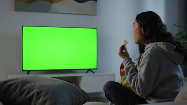Green Screen woman looking at horizontal on television while sitting sofa eating chips young adult watching isolated background with chroma key and mockup template in living room after work scene 1