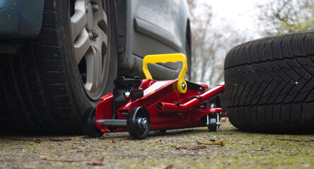 Red Hydraulic Floor Jack Under Car With Yellow Handle Beside Spare Tire On Ground
