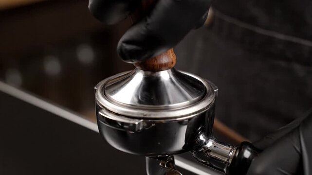 Close-up of a barista tamping espresso grounds with a steel tool. Black gloves are visible