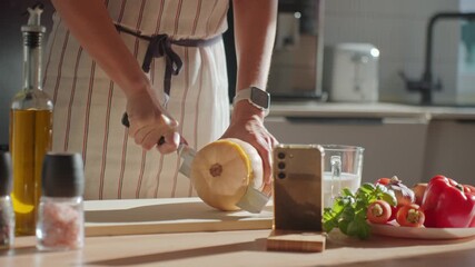 Person in apron cutting butternut squash on wooden board in kitchen. Close-up of woman slicing fresh pumpkin with fresh vegetables and herbs on table. Concept of healthy eating and home cooking