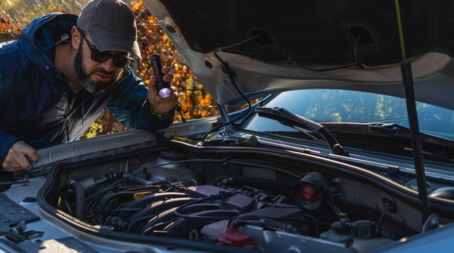 Man inspects car engine with flashlight in sunny outdoor setting