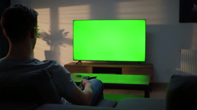Green Screen man watching television in living room evening young guy sitting on sofa against tv with chroma back view male person switching channels using remote controller scene 3 distortion