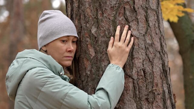 Melancholic woman wearing a beanie and jacket seeking solace by embracing a large pine tree trunk, finding comfort and connection with nature in an atmospheric autumn forest during a difficult time