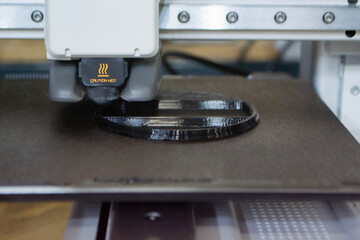 3D printer close up. Working 3D printer in process of printing object from molten black plastic. 3D printer creating model by flowing liquid plastic from extruder of printer. 3D printing technology
