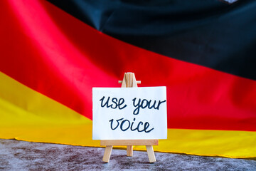 Paper note with words USE YOUR VOICE on National symbol of Germany. German wave silky flag...