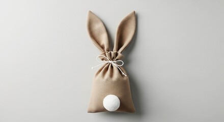 Obraz premium A cute Easter bunny gift bag made of beige fabric with a white pom-pom tail on a gray background.