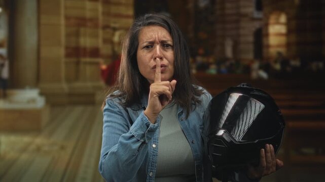Middle age woman holding a glossy black motorcycle helmet, finger to lips for silence while standing inside a church nave with wooden pews and stone columns; quiet contemplation.