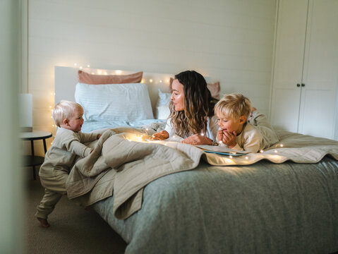 Mum and son lying on their belly and toddler baby climbing on the bed on the bed with string lights