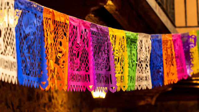 A colorful string of paper lanterns hang from the ceiling. The lanterns are of different colors. Mexican Day of the Dead tradition, offerings, food, colors, decorations, and papel picado, La Catrina.