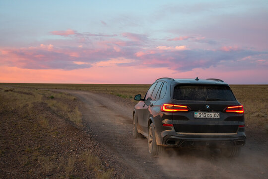Naryn, Kyrgyzstan - May 13, 2024: Rear view of a dark colored BMW X5 G05 SUV driving on a dirt track and kicking up a cloud of dust against a vibrant pink and orange sunset sky