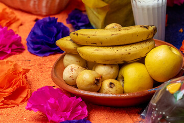 A bowl of fruit with bananas, apples, and oranges on a table. The bowl is on a table with a pink. Mexican Day of the Dead tradition, offerings, food, colors, decorations, and papel picado, La Catrina.
