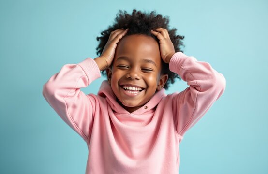 Excited african american boy laughs holding hands on head. Happy child joyfully posing in studio with closed eyes. Afro kid in pink hoodie smiles broadly at camera on cyan backdrop. - Powered by Adobe
