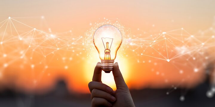 Hand holding a lit incandescent light bulb against a sunset with network connections