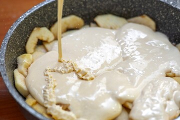 Thin stream of liquid batter being poured over apple layer in a pan for making pie
