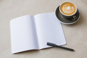 Blank open notebook, pen and a cup of cappuccino with a heart-shaped pattern on the foam.