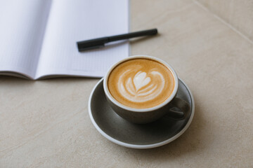 A cappuccino with a heart-shaped pattern on the foam and an open blank notebook with a pen on...