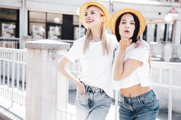 Two young beautiful smiling hipster female in trendy summer white t-shirt and jeans clothes. Carefree women posing in street. Positive models having fun. Cheerful and happy. In hat. Give air kiss