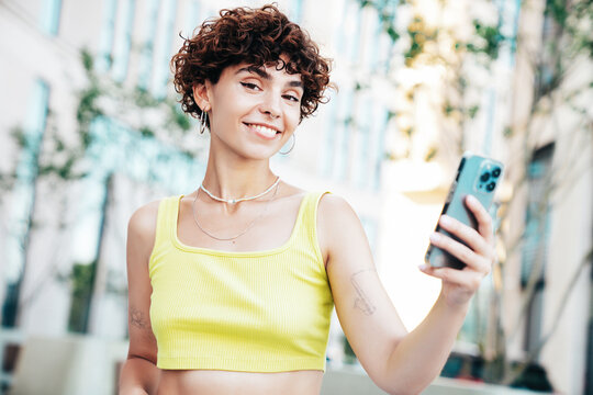 Young beautiful smiling hipster woman in trendy summer clothes. Carefree woman with curls hairstyle, in the street. Positive model holds mobile phone, looks at cellphone screen, uses smartphone apps