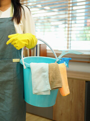 A cleaning woman is standing inside a building holding a bucket fulfilled with chemicals and facilities for tidying.