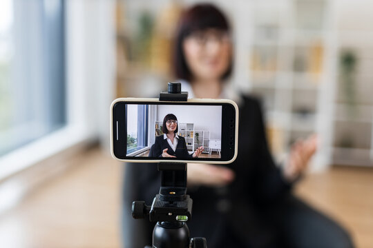 Caucasian woman recording video blog about business topics using smartphone camera on tripod. Professional indoor office setup emphasizes modern technology, content creation, and professionalism.