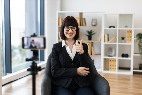 Caucasian woman in her 30s recording business-related video blog using smartphone at home office, wearing professional suit and thumbs up gesture. Bright environment with shelves and modern office.
