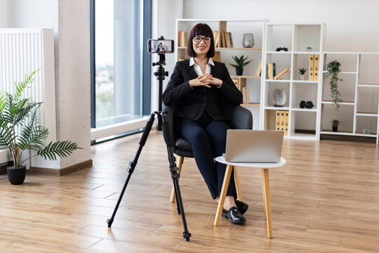 Caucasian woman aged 30-40 recording business vlog through camera in modern office. Professional attire shows corporate setting. Bright room features creative workspace environment.