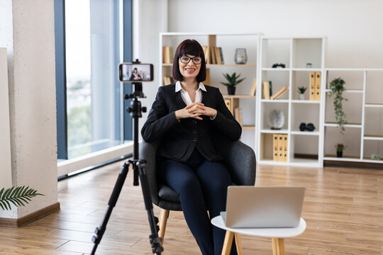 Caucasian adult woman using camera for recording business-related content in modern office environment. Focus on creating professional material remotely with digital tools.