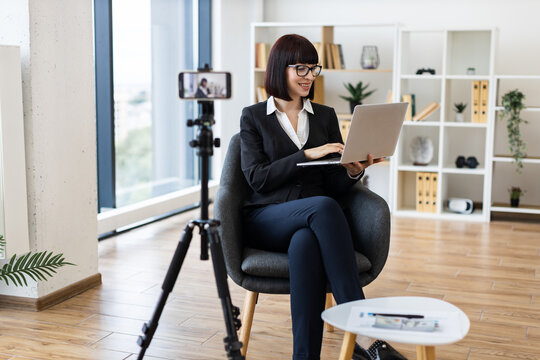 Young Caucasian woman in formal attire recording professional blog content on camera in spacious office environment, smiling while using laptop, showcasing modern working setting involving technology