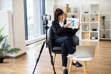 Caucasian woman presenting business analytics on camera through blogging using report and statistics chart.