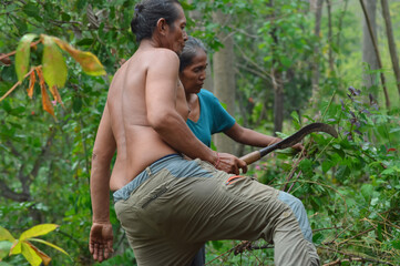 A shirtless father steps through brush carrying a sickle while his wife gathers weeds behind him in their dense tropical farmland filled with green plants and shrubs.