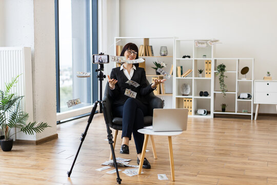 Caucasian woman in business attire sitting in modern office, blogging about investments on camera while confidently tossing dollar bills, showcasing entrepreneurial success, positivity.