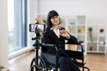 Caucasian woman in business attire sits in wheelchair recording informative business blog on smartphone. Modern interior, professional appearance, creating content for business communication.