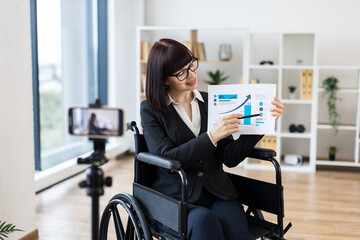 Caucasian woman in wheelchair holding chart showing business data while blogging on camera