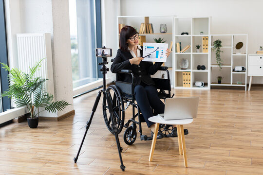 Caucasian woman in wheelchair creates business presentation, showcasing statistics and data on camera. Professional environment.