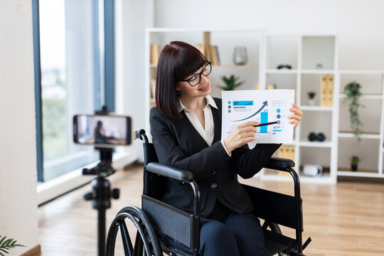 Caucasian woman in wheelchair holding chart showing business data while blogging on camera