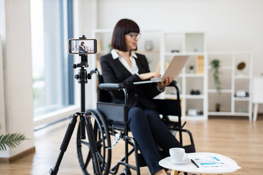 Caucasian woman in wheelchair using laptop, creating business content with phone camera, workplace setting