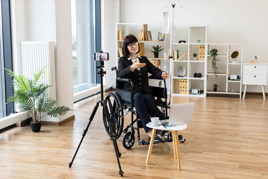 Caucasian woman recording blog with phone while working from wheelchair and receiving IV treatment. Modern office space, smart devices, professional attire, and healthcare equipment