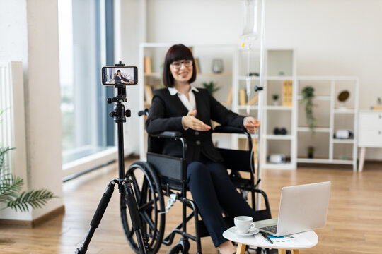 Caucasian woman in wheelchair receiving intravenous drip recording vlog about combining work with treatment using smartphone camera and laptop in modern office space