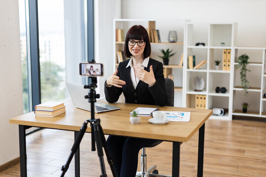 Caucasian woman in her 30s sitting at desk in modern office recording video blog about business and communication, professional setting and educational content, emphasizing workplace skills.
