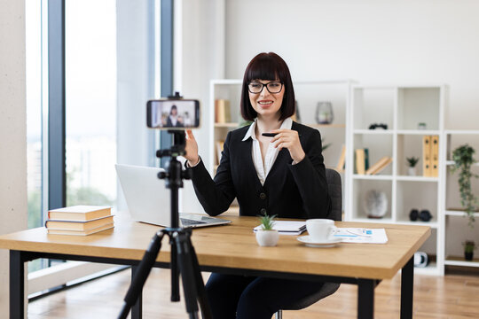 Caucasian woman in her 30s sitting at desk in modern office recording video blog about business and communication, professional setting and educational content, emphasizing workplace skills.