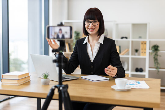 Caucasian adult woman wearing professional attire recording video blog about business topics using smartphone camera in modern office environment. Includes elements like notebook, laptop. - Powered by Adobe
