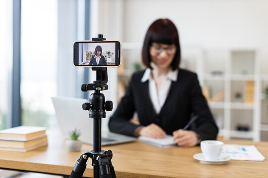 Caucasian woman wearing black suit recording video blog in office environment using smartphone on tripod