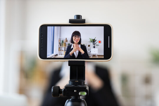 Caucasian adult woman sitting at desk, recording business blog with smartphone camera. Image shows professional woman in modern office environment creating content about business topics.