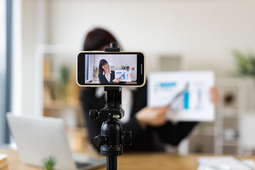 Caucasian woman recording business blog using smartphone on tripod in office environment. Discussing chart data in video. Professional creating online content. Relates to business, content creation.