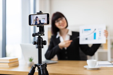 Caucasian woman recording business blog using smartphone on tripod in office environment. Discussing chart data in video. Professional creating online content. Relates to business, content creation.
