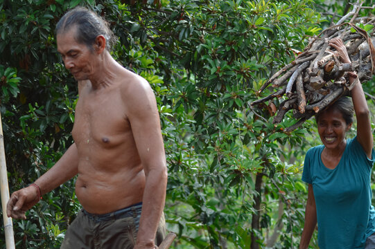 A cheerful middle-aged wife smiles proudly after reaching the top of the hill, balancing a heavy bundle of firewood on her head, while her shirtless husband walks beside her through sunny farmland.