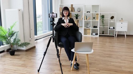 Caucasian woman recording video blog on business topics sitting in stylish office interior. Professional setup includes camera, tripod, laptop demonstrating work focus and modern office aesthetic. - Powered by Adobe