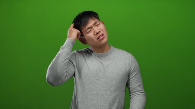 Young man with confused expression scratching head against green background, wearing casual gray sweater and isolated over vivid wall