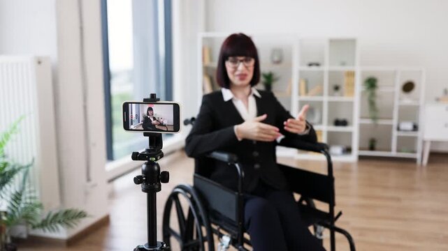 Caucasian woman wearing business attire sits in wheelchair recording video blog in office using smartphone. Professional setting conveys ideas of inclusivity accessibility business presentation
