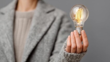 A person in a grey sweater holds a glowing incandescent light bulb against a grey background